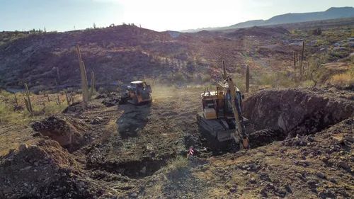 Construction site in desert. Excavator digs, truck parked in pit. Mountains in background.