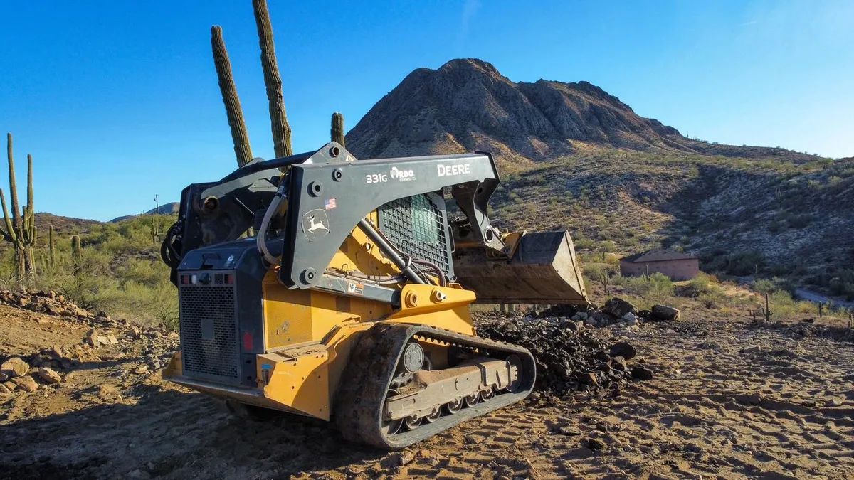 Yellow skid steer loader in desert landscape, scooping dirt. Mountain in background, blue sky.