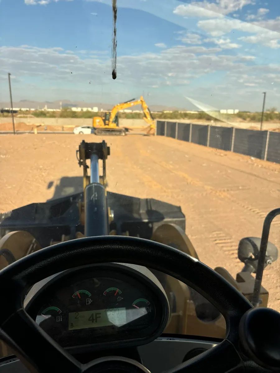 View from inside a loader at a construction site, with an excavator in the distance. Sunny, open space.