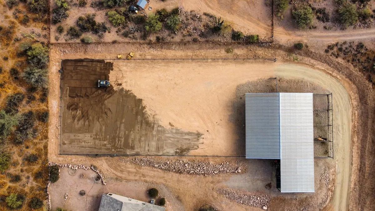 Aerial view: Dirt area being graded by a machine next to a white-roofed building and a gravel driveway in a desert landscape.