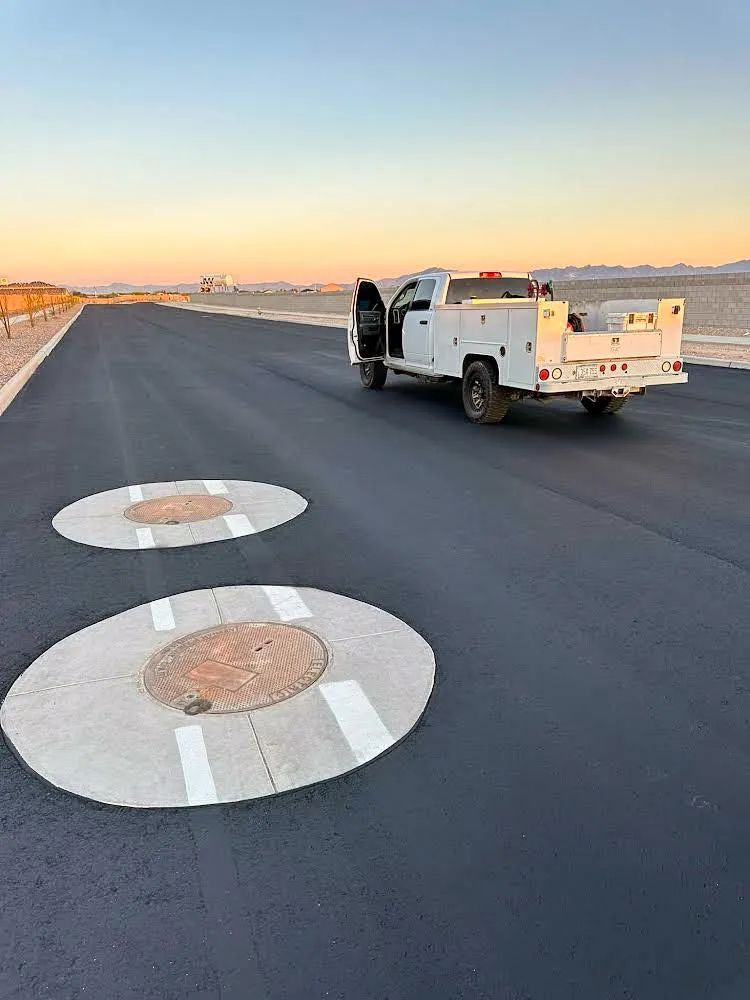 A utility truck parked on newly paved road, two manhole covers in foreground, sky at sunset.