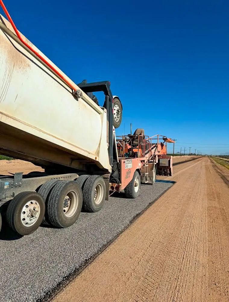 Dump truck unloading gravel onto a new road, against a bright blue sky.