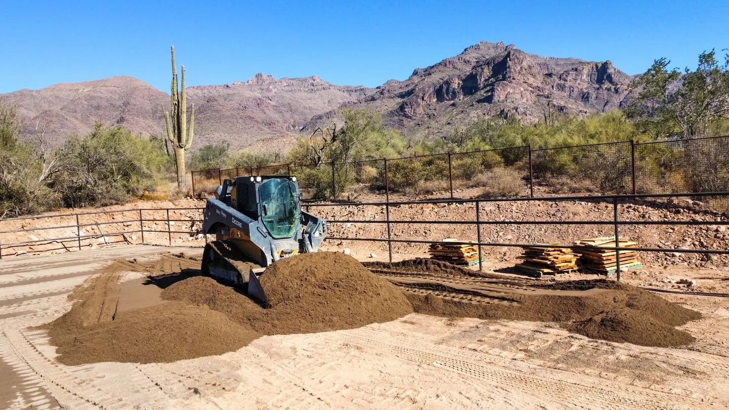 Skid steer moving soil near a fence, with mountains and cacti in the background.