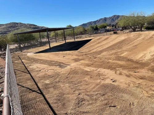 Dirt arena with a shaded structure, fence, and mountain backdrop under a blue sky.