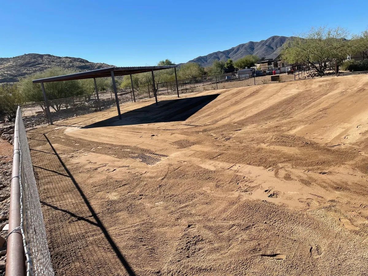 Dirt area with a covered structure, trees, and a fence against a mountainous backdrop on a sunny day.