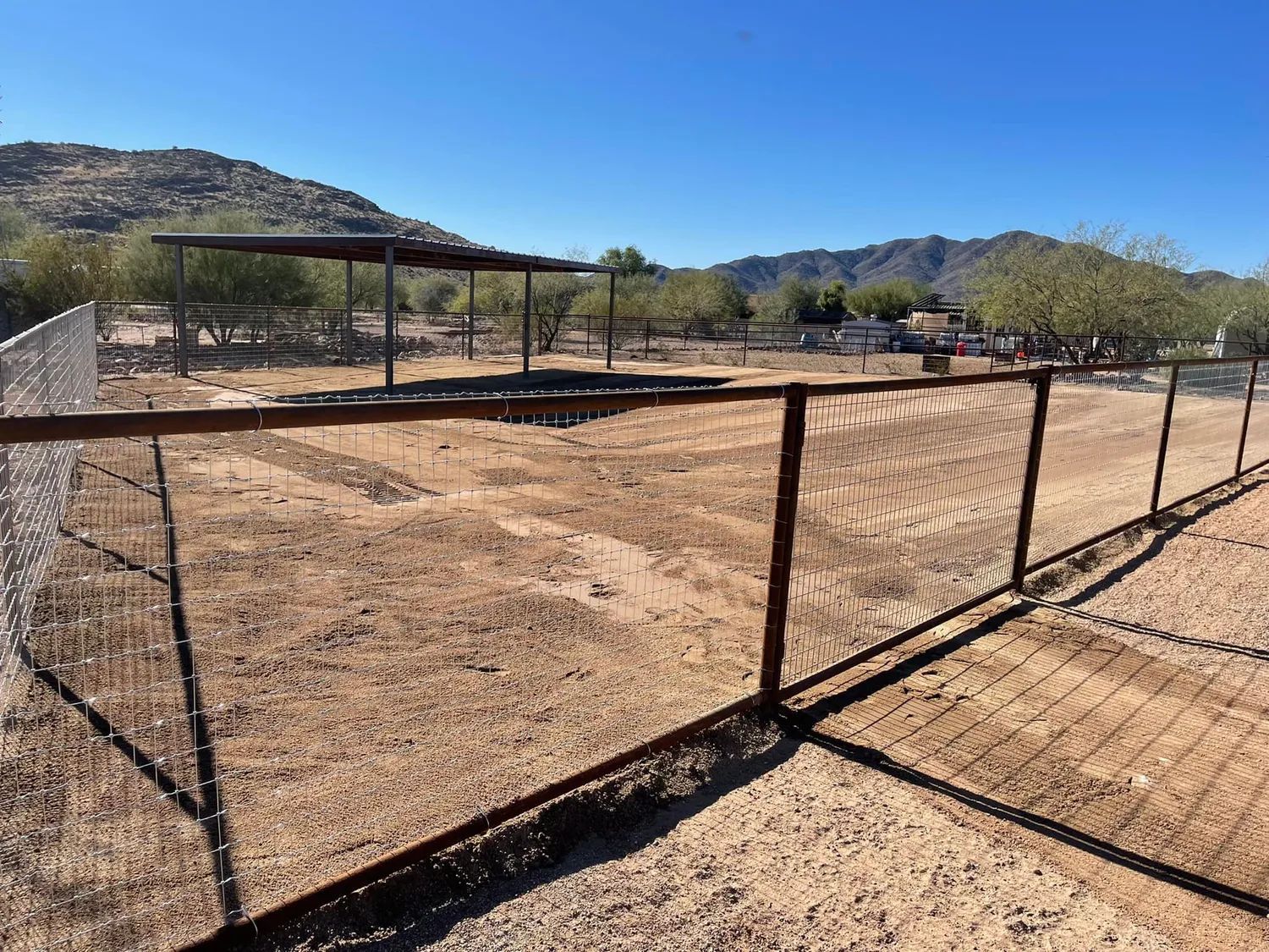 Fenced, dirt-covered outdoor arena under a blue sky. A large shelter is in the distance.