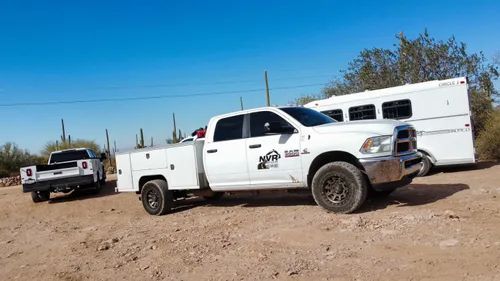 White work truck with trailer parked on dirt road in sunny desert. Another truck behind.