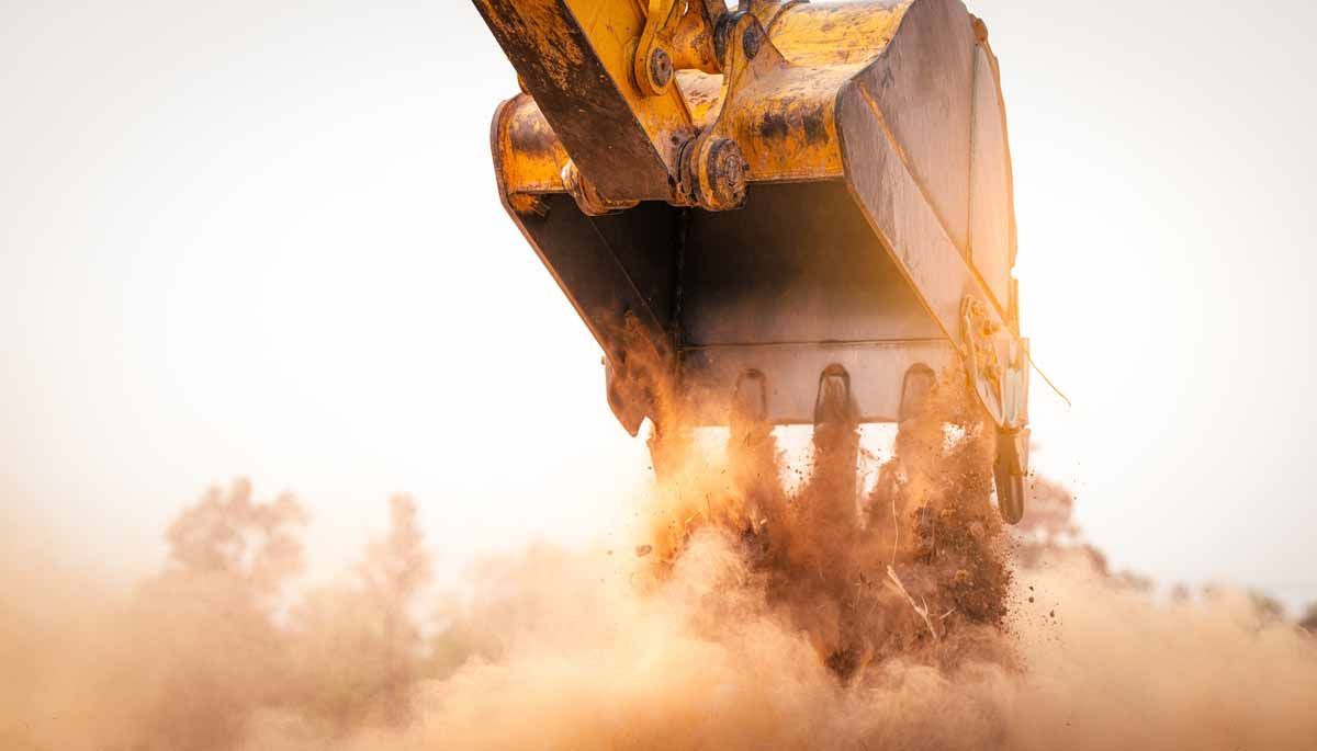 Yellow excavator bucket digging, kicking up dust into the air, setting sun.