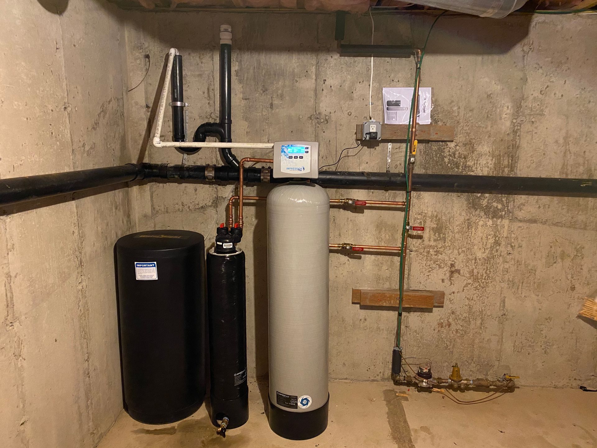 Water treatment system in a basement. Includes tanks, pipes, and control panel against a concrete wall.