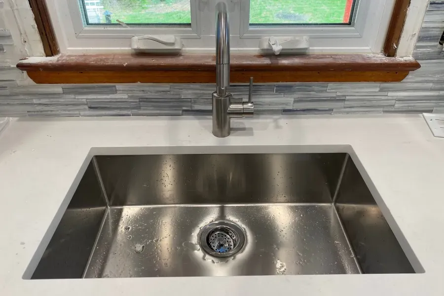 Stainless steel kitchen sink with faucet, white countertop, and window in the background.