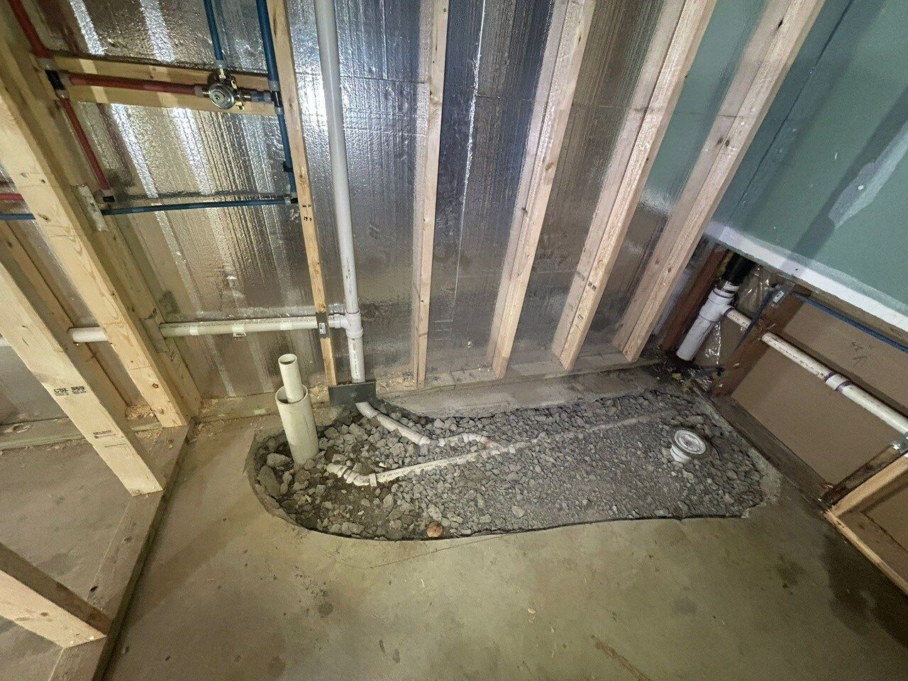 Framed wall with plumbing and gravel area on concrete floor; construction in progress.