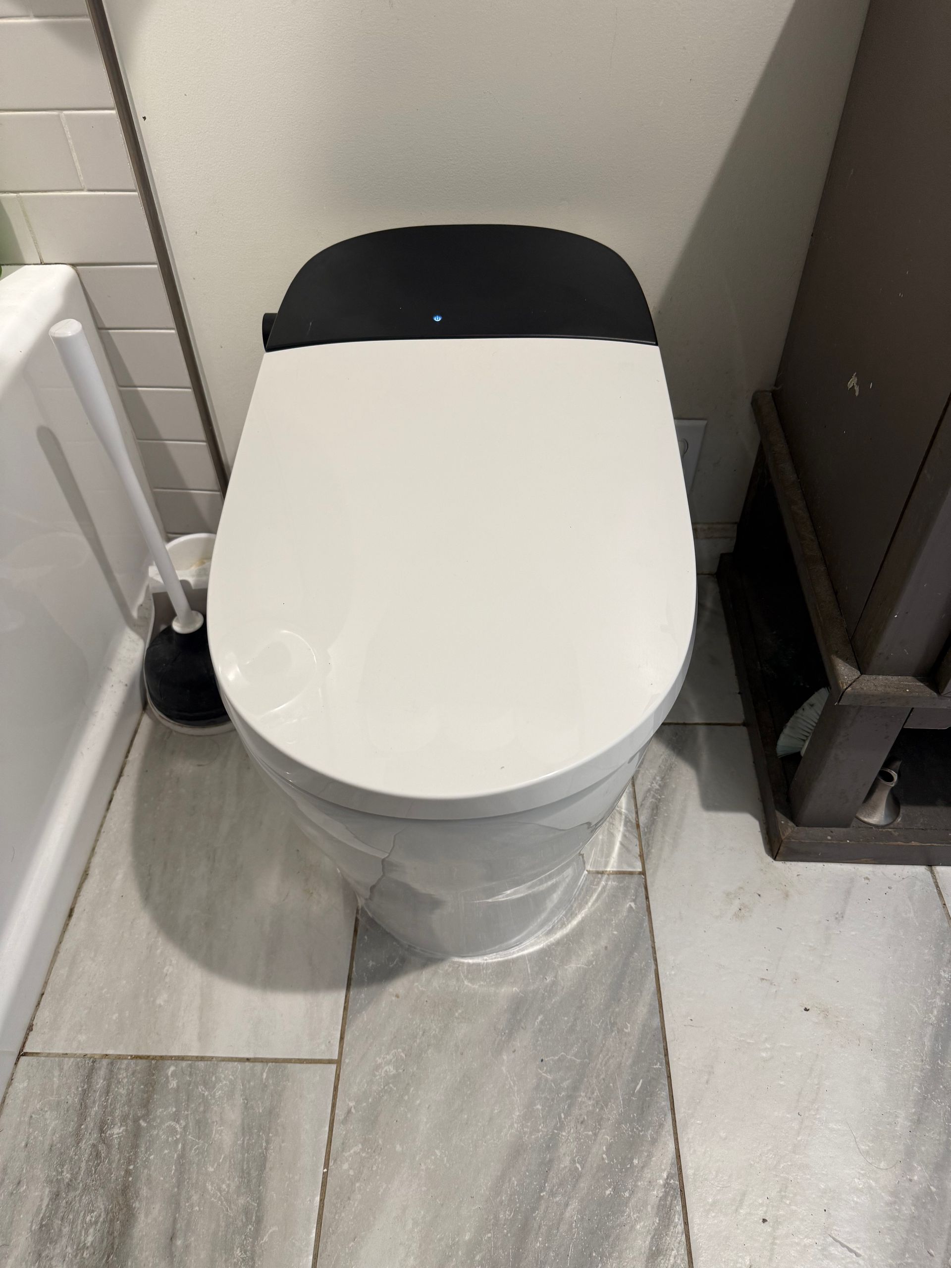 White and black toilet in a bathroom, with a brush to the left and a cabinet to the right.