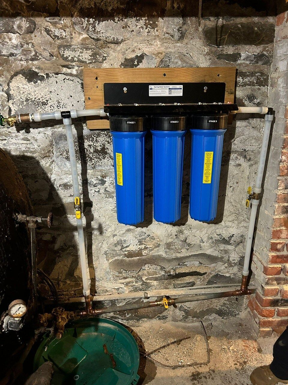 Three blue water filters attached to a wooden board on a basement wall with pipes and valves.