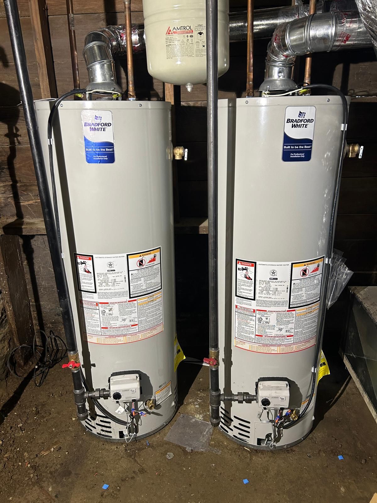 Two upright water heaters side by side, silver color, in a basement setting.