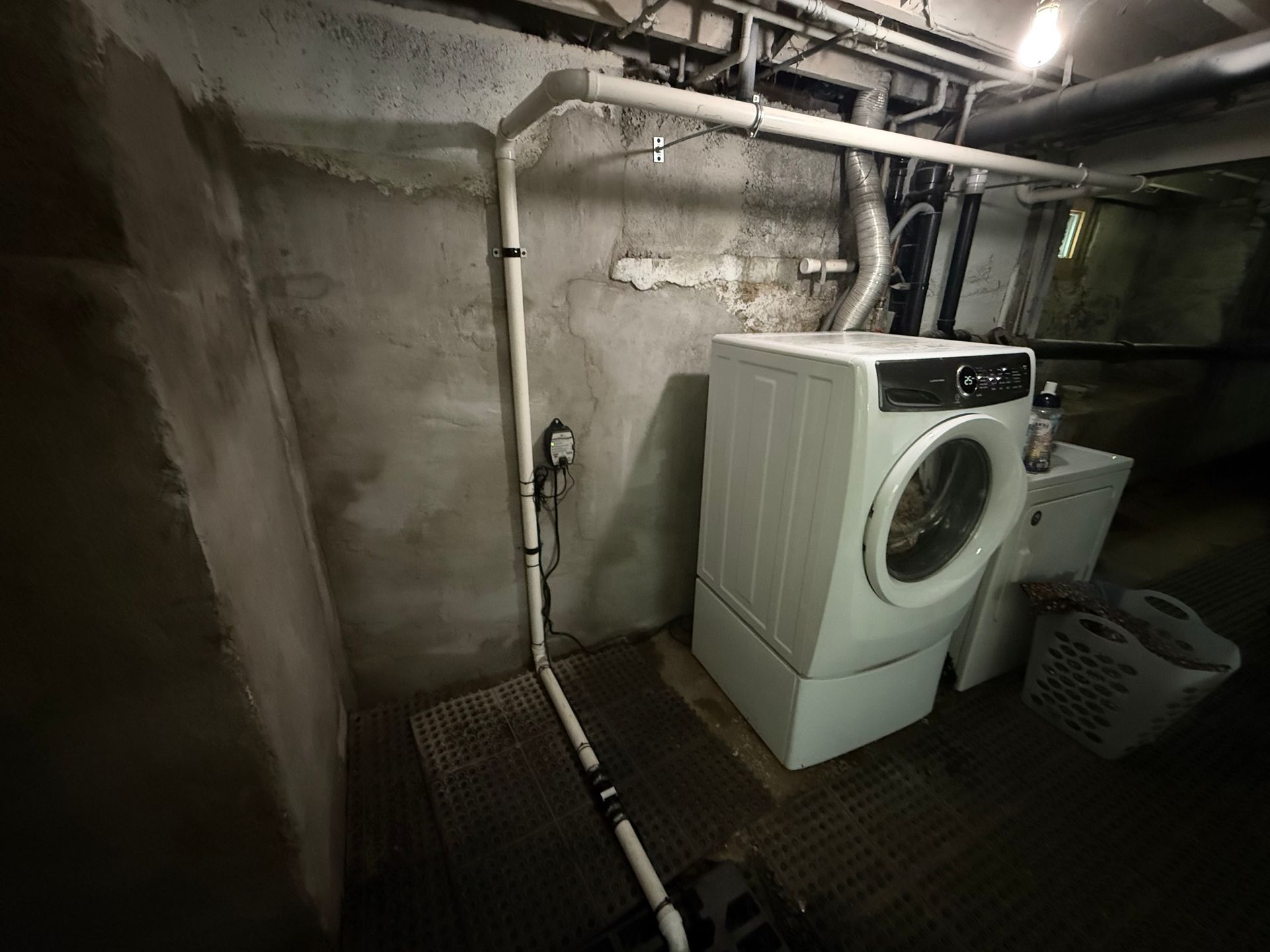 A basement laundry area with a washing machine, pipes, and a laundry basket on a metal grate.