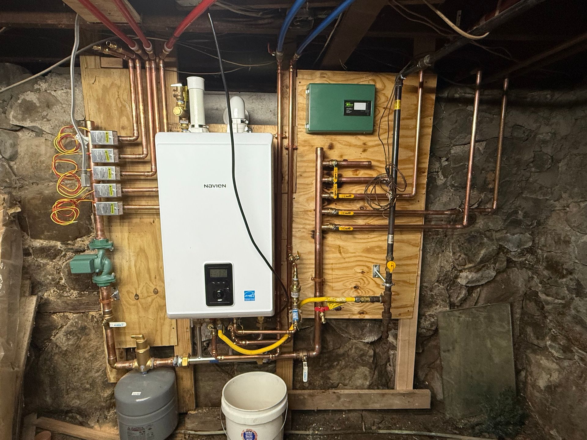 A boiler system installed in a basement, with copper pipes, a white boiler, and a green control panel.