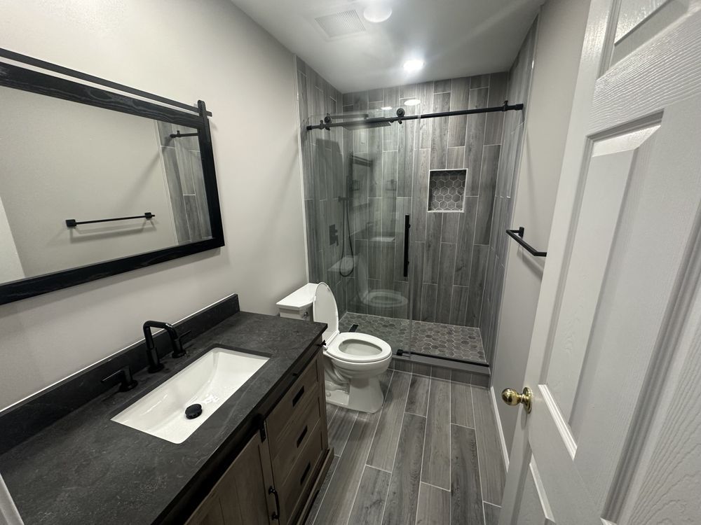 Modern bathroom with gray tile shower, wood vanity, and a black-framed mirror.
