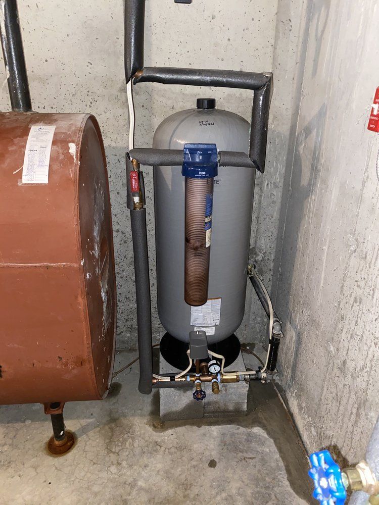 A tall, gray water tank with pipes and a filter, set in a basement next to an oil tank.