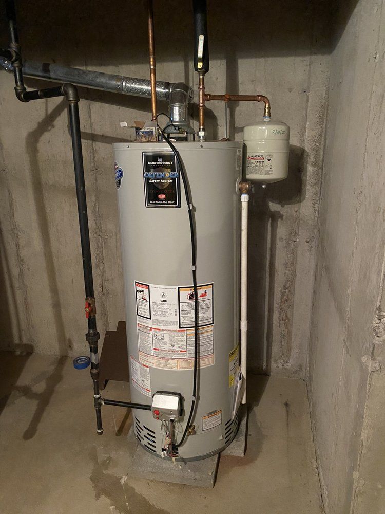 Gray water heater in a concrete-walled room, with pipes and an expansion tank attached.