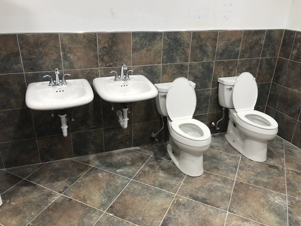 Two sinks and two toilets in a bathroom with brown tile flooring and walls.