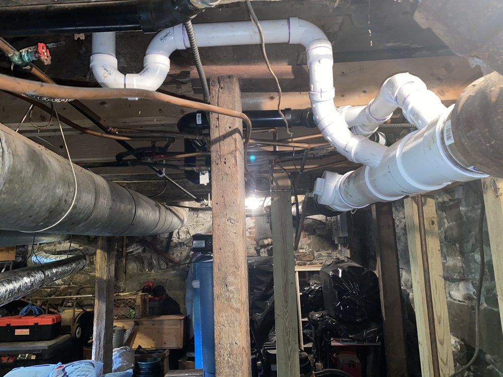 Underneath a wooden structure, white PVC pipes are connected to existing pipes; a basement setting.