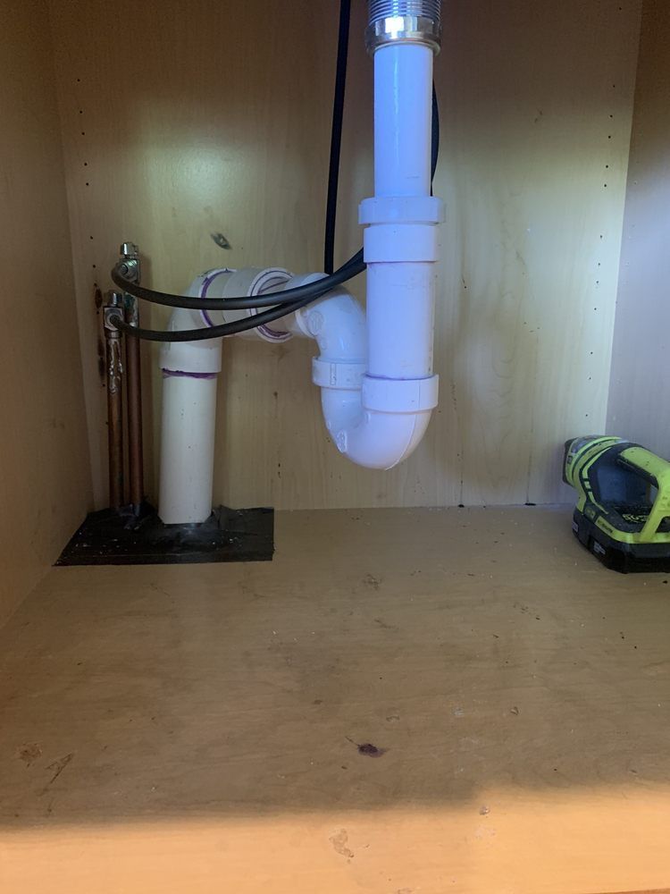 White plastic sink plumbing with a P-trap inside a wooden cabinet. A cordless drill is visible.