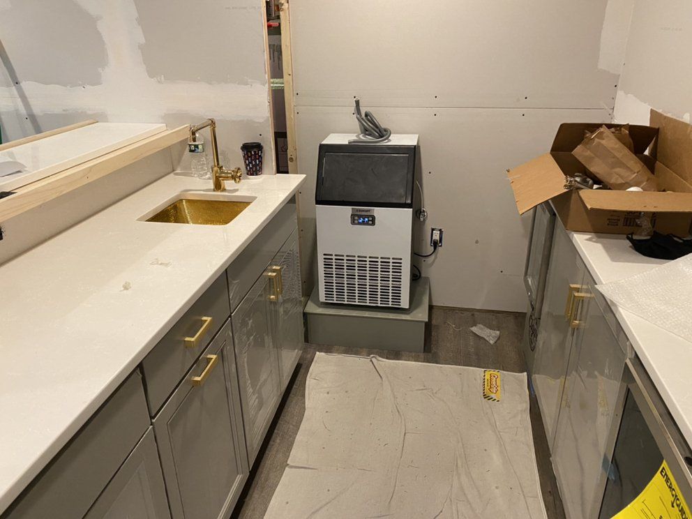 Gray kitchen cabinets with gold hardware and a white countertop. A gold faucet, sink, and ice maker are present.