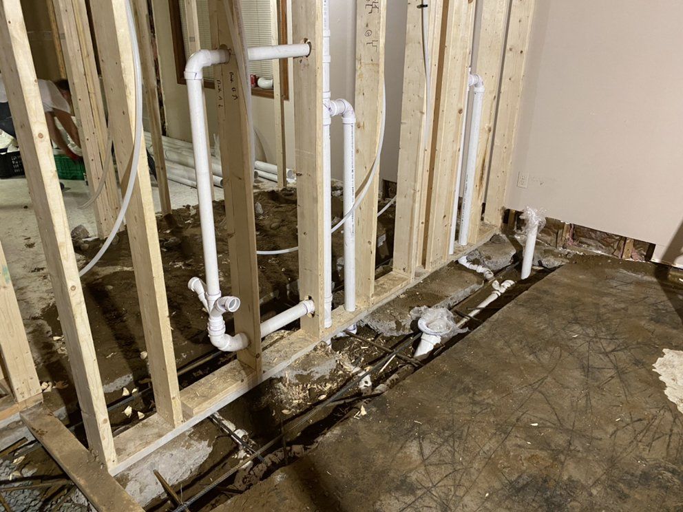 Interior view of a bathroom remodel in progress with exposed white PVC pipes against wooden wall studs.