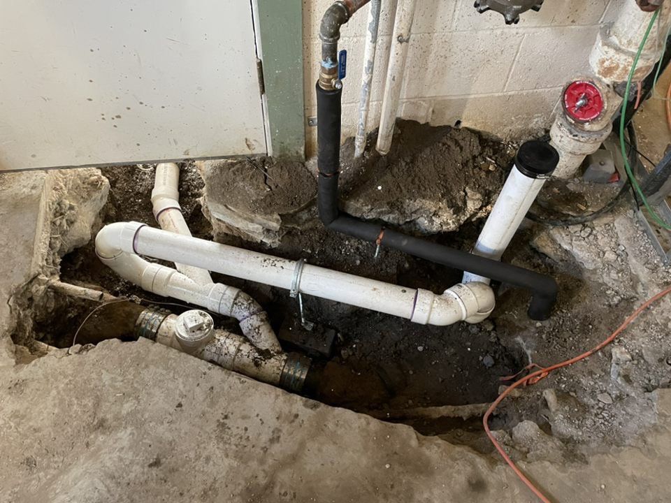 Exposed plumbing pipes in a concrete floor excavation. White PVC pipes and black insulated pipes are visible.