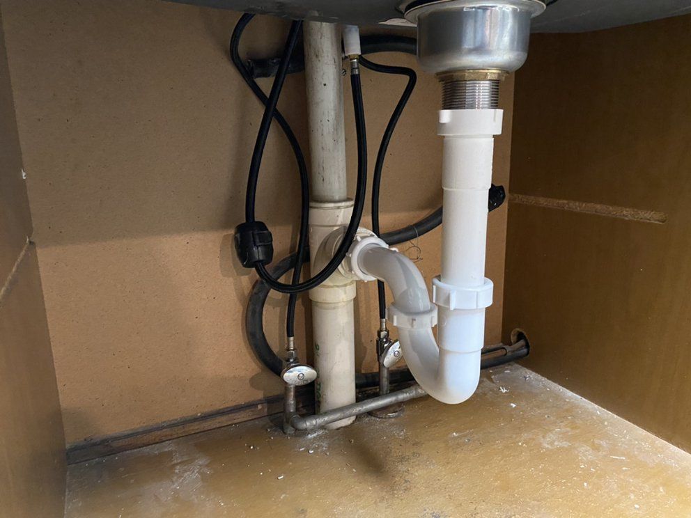 Underside of a sink showing white plumbing pipes and black water supply lines inside a beige cabinet.