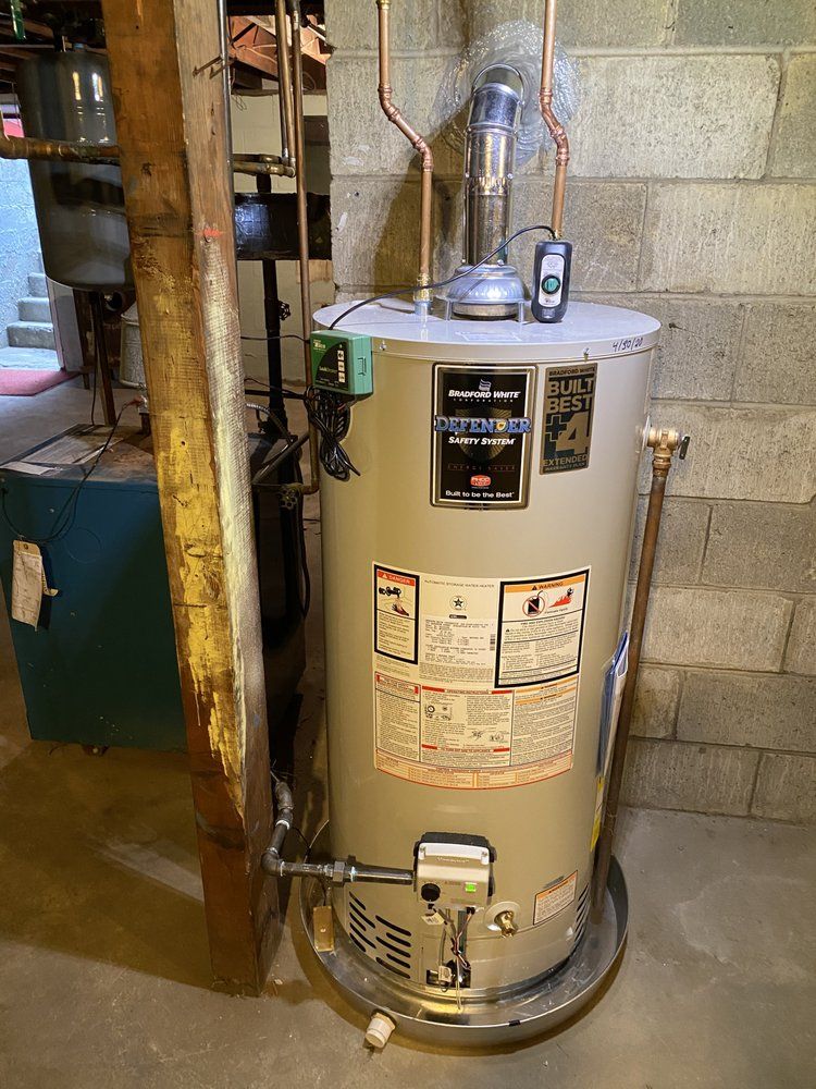 A water heater in a basement. It's cylindrical, light-colored with visible controls, a flue, and pipes against a concrete block wall.