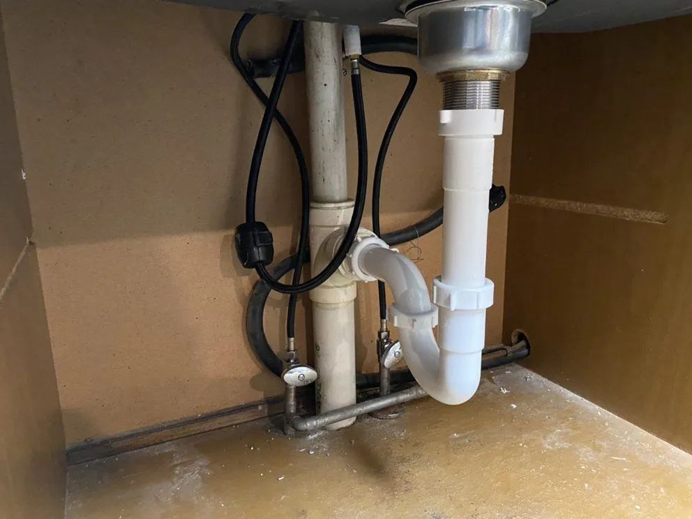 Under-sink plumbing: white pipes, chrome drain, black hoses, cabinet interior.