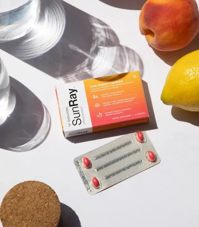 Caja y blíster de suplemento SunRay, cápsulas rojas, con melocotón, limón, corcho y agua sobre superficie blanca.