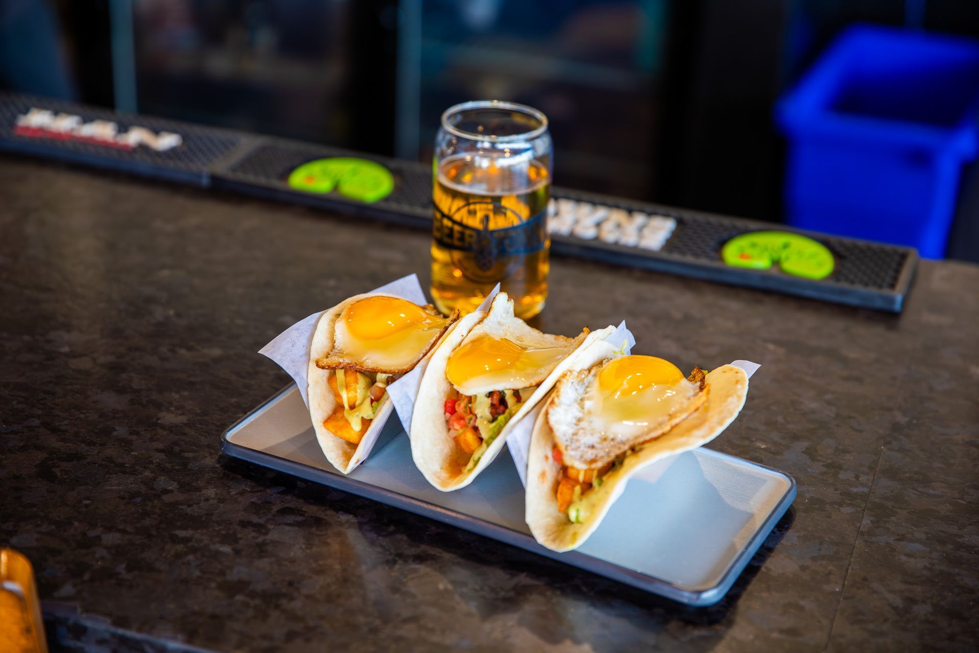 A close up of three tacos on a plate with a glass of beer in the background.