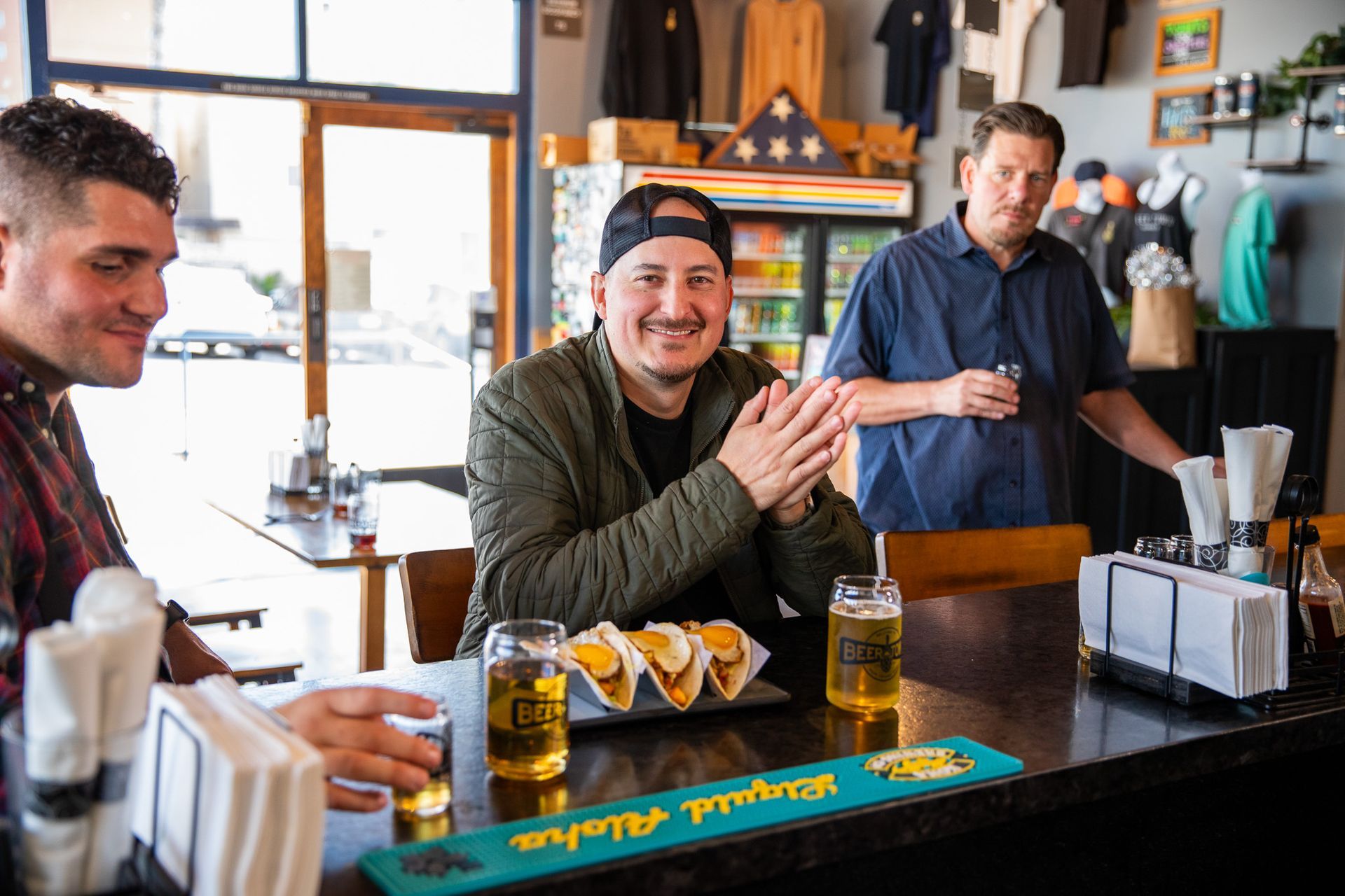 Three men are sitting at a bar drinking beer and eating tacos.