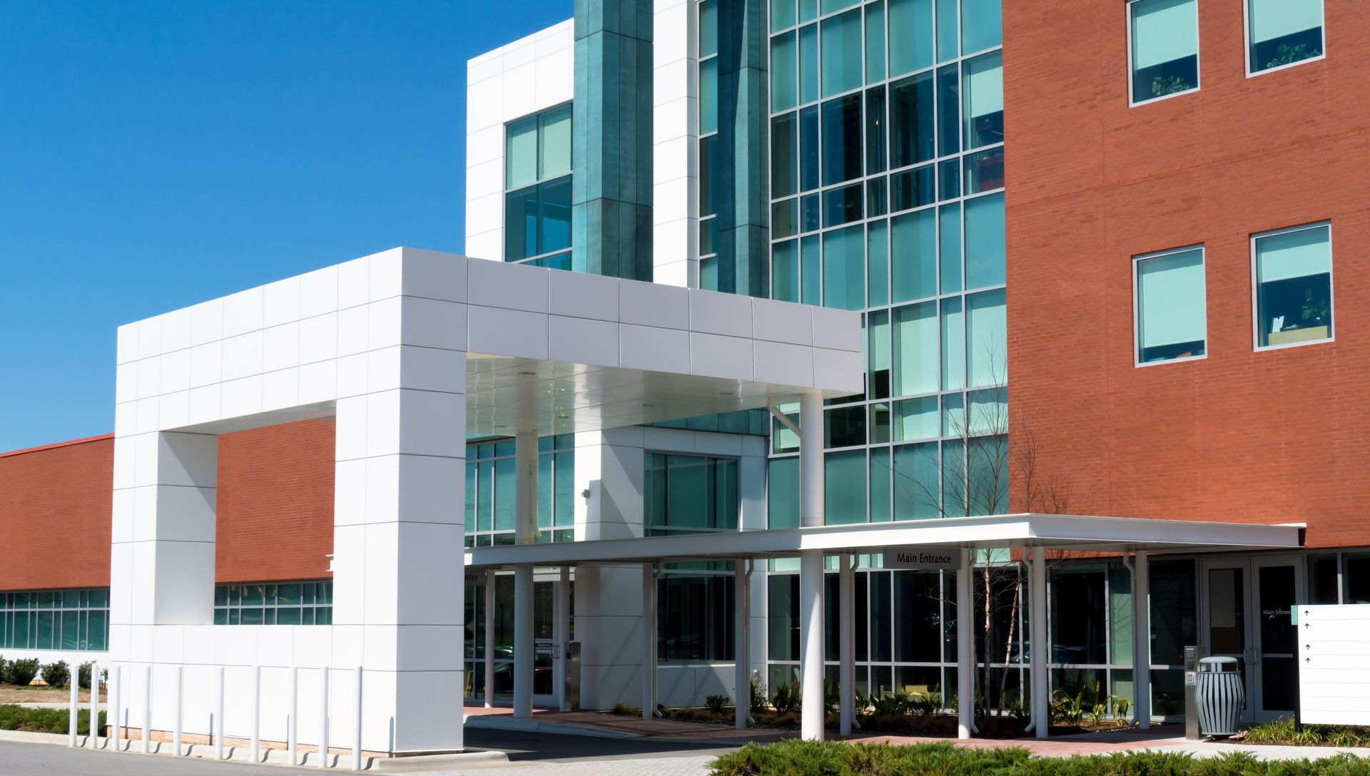 Modern building entrance with white canopy, glass windows, and red brick facade against a blue sky.