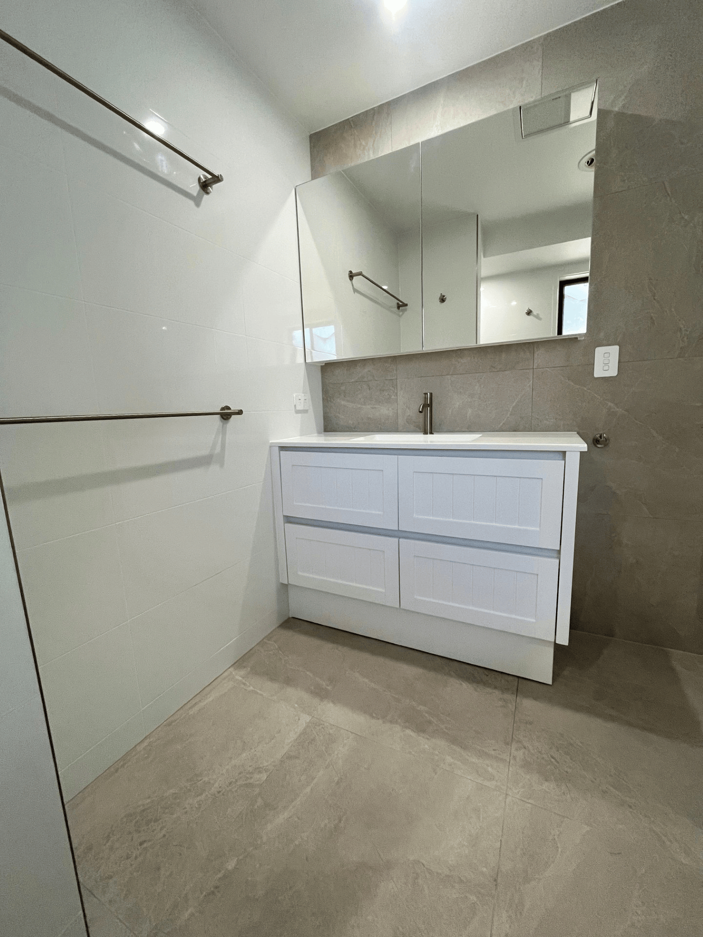A modern bathroom with a white vanity, a large rectangular mirror, and beige tiled walls and floors.