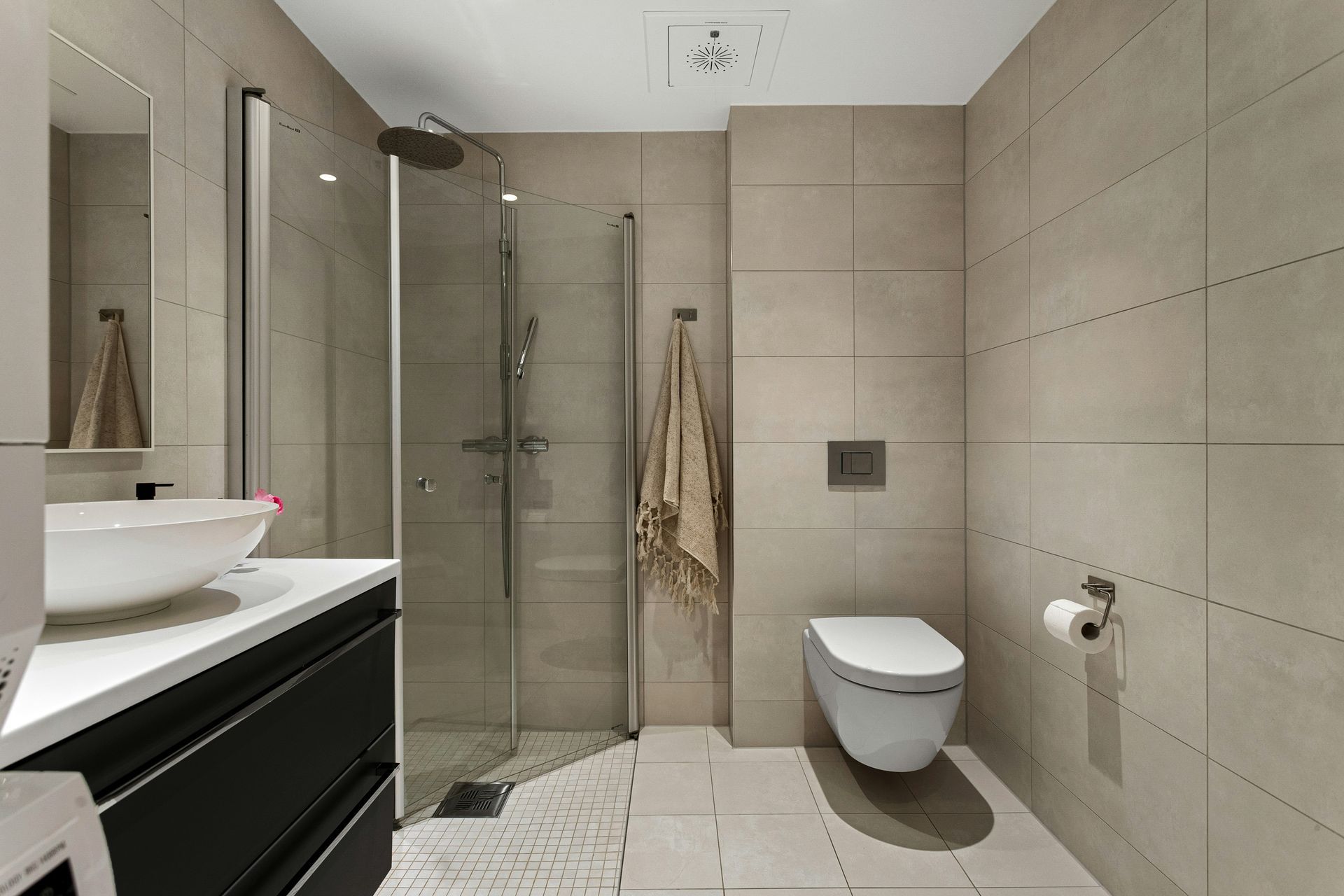 A modern bathroom with light-toned tiled walls, a black vanity, a white vessel sink, a walk-in shower, and a wall-hung toilet.