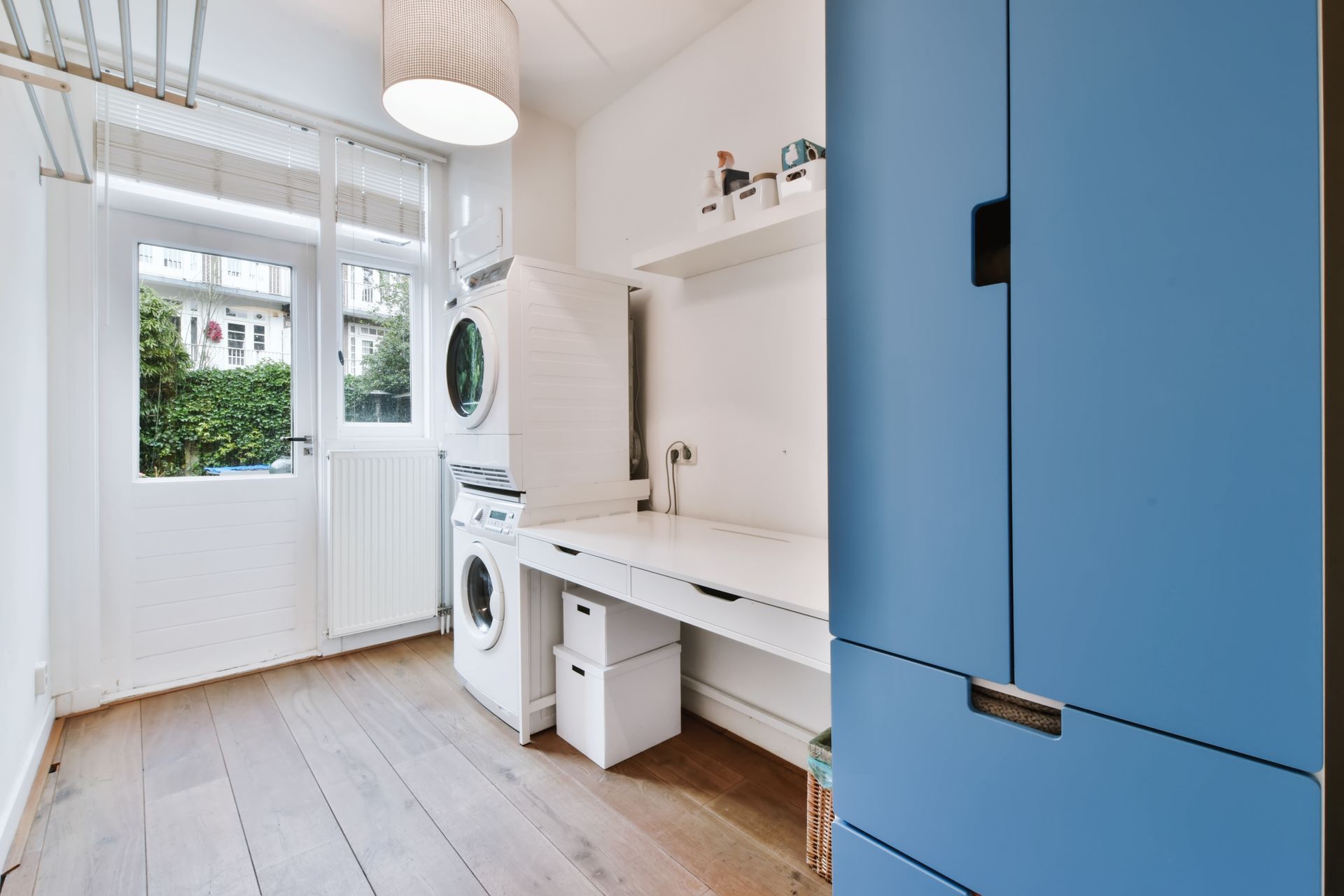 A modern laundry room with a stacked washer and dryer, white work counter, storage boxes, and a tall blue cabinet.