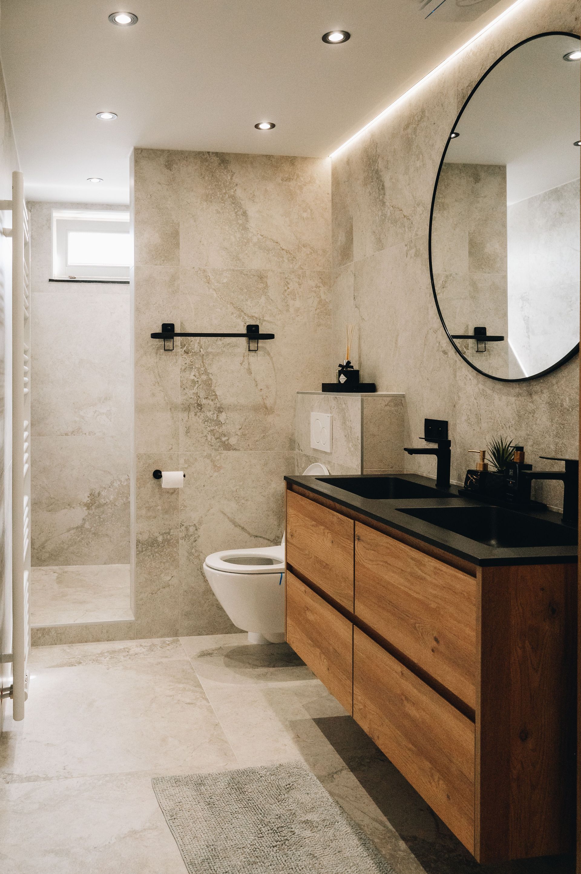 Modern bathroom featuring a wood vanity with a black countertop, oval mirror, walk-in shower, and stone-tiled walls.