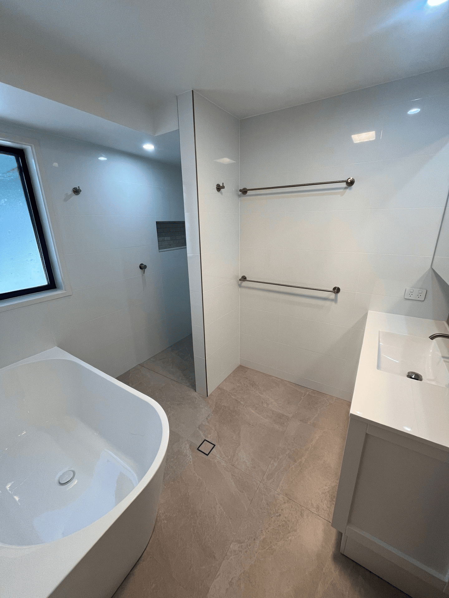 Modern bathroom with a white freestanding tub, vanity, wall-mounted towel racks, and beige tile flooring.