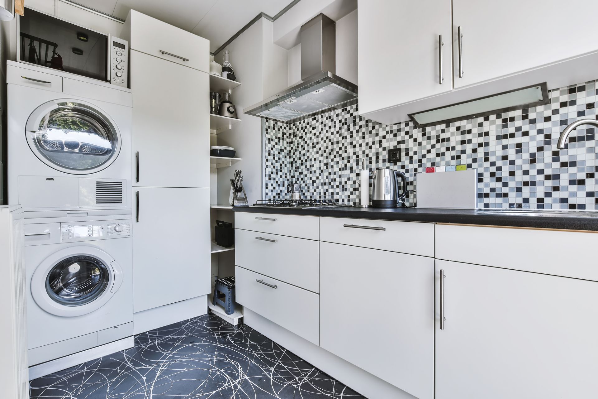A modern kitchen with white cabinets, black and white mosaic tile backsplash, a stacked washer and dryer, and dark floors.