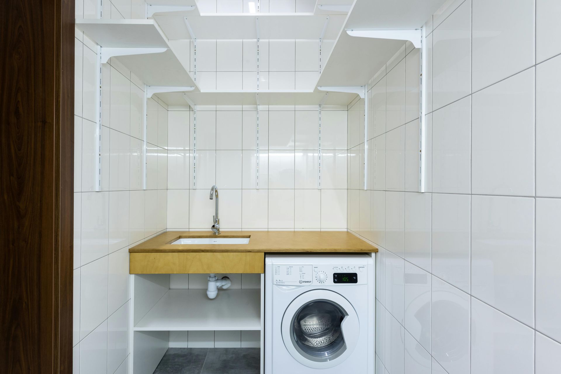 A laundry area featuring a white washing machine, a sink with a wooden countertop, and open white shelving against white tiles.