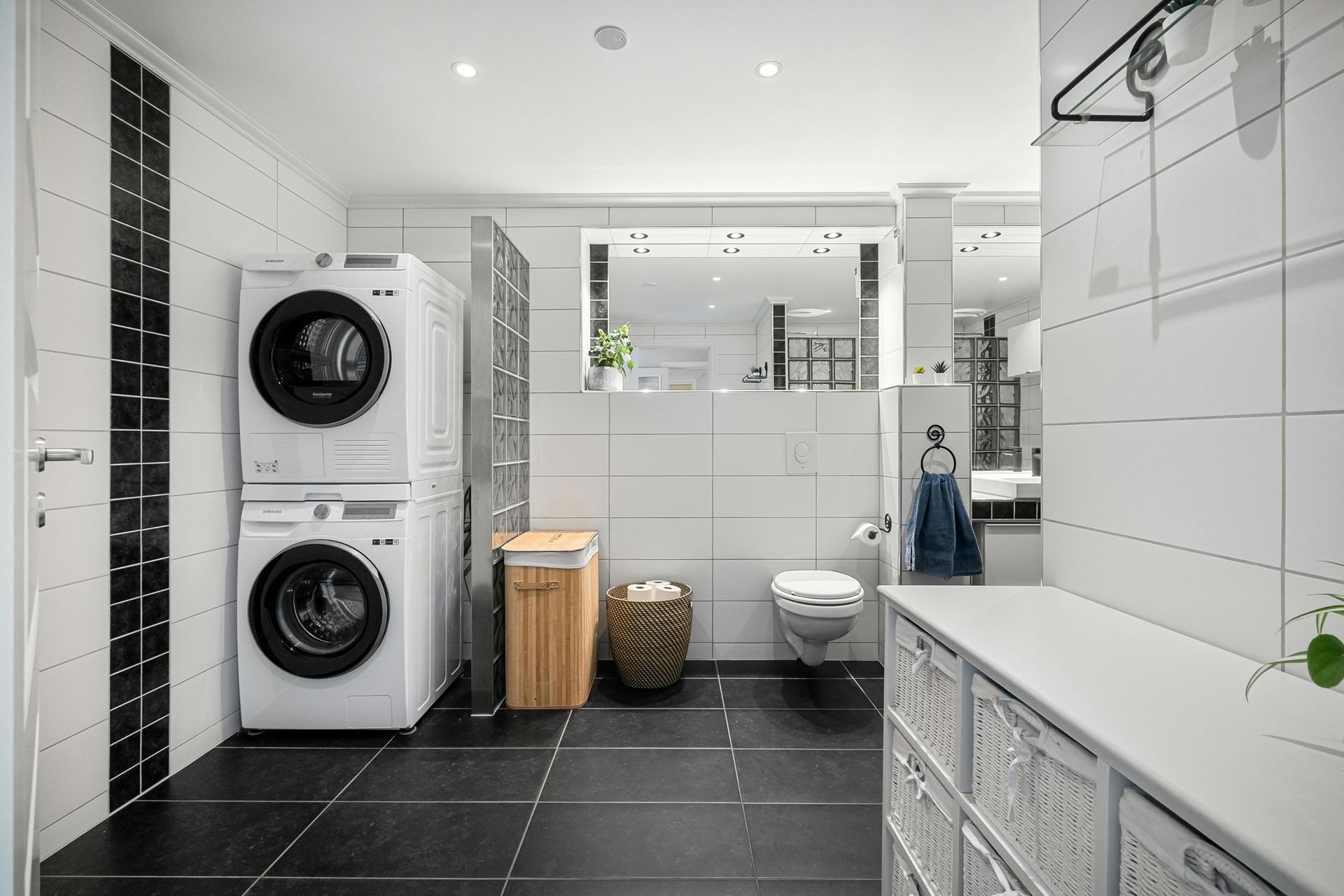 A modern laundry room and bathroom with white tiled walls, black floor tiles, a stacked washer-dryer, and a toilet.