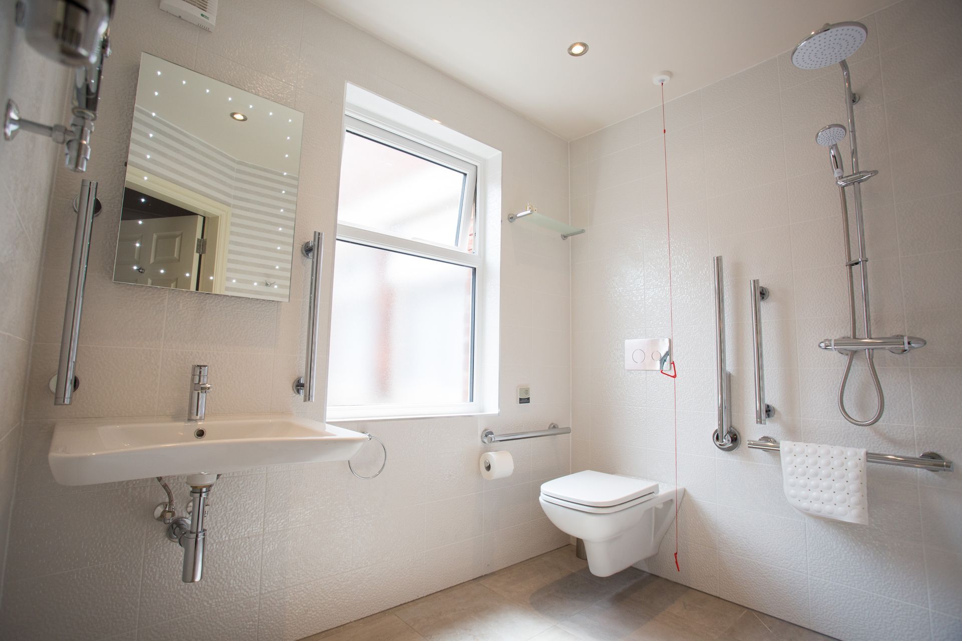 A brightly lit, accessible white bathroom with a wall-mounted toilet, sink, chrome safety grab bars, and a walk-in shower.