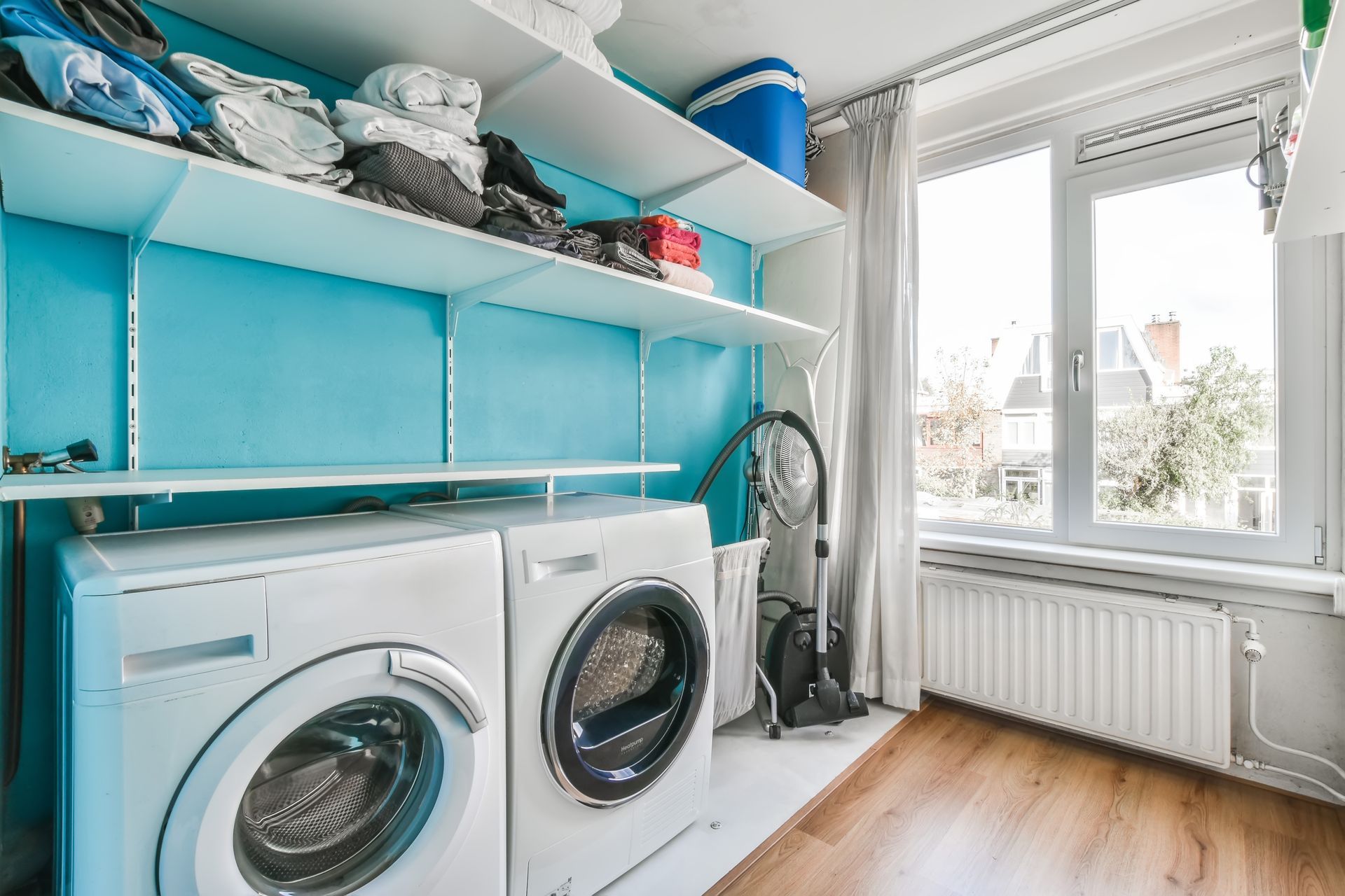 Laundry room with two white washing machines against a bright blue wall, overhead shelving, and a nearby window.
