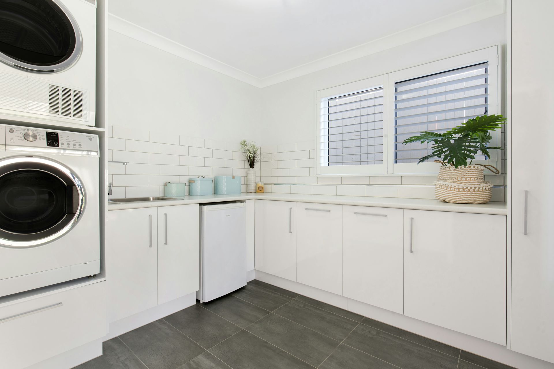 A bright white laundry room featuring stacked washers, glossy cabinets, a small refrigerator, and a potted plant.