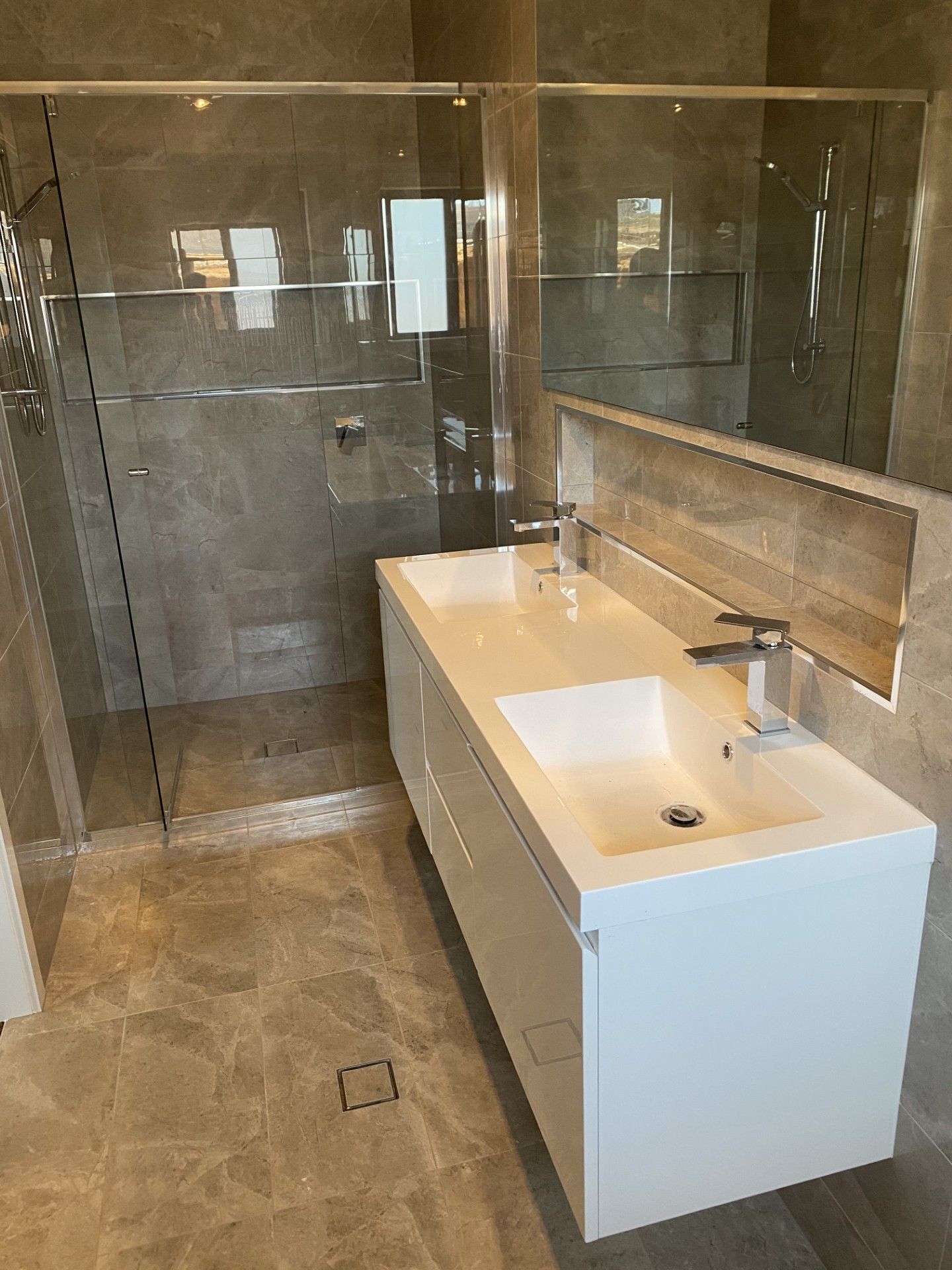 Modern bathroom with a double white vanity, silver faucets, and a glass-enclosed walk-in shower with grey stone tiles.
