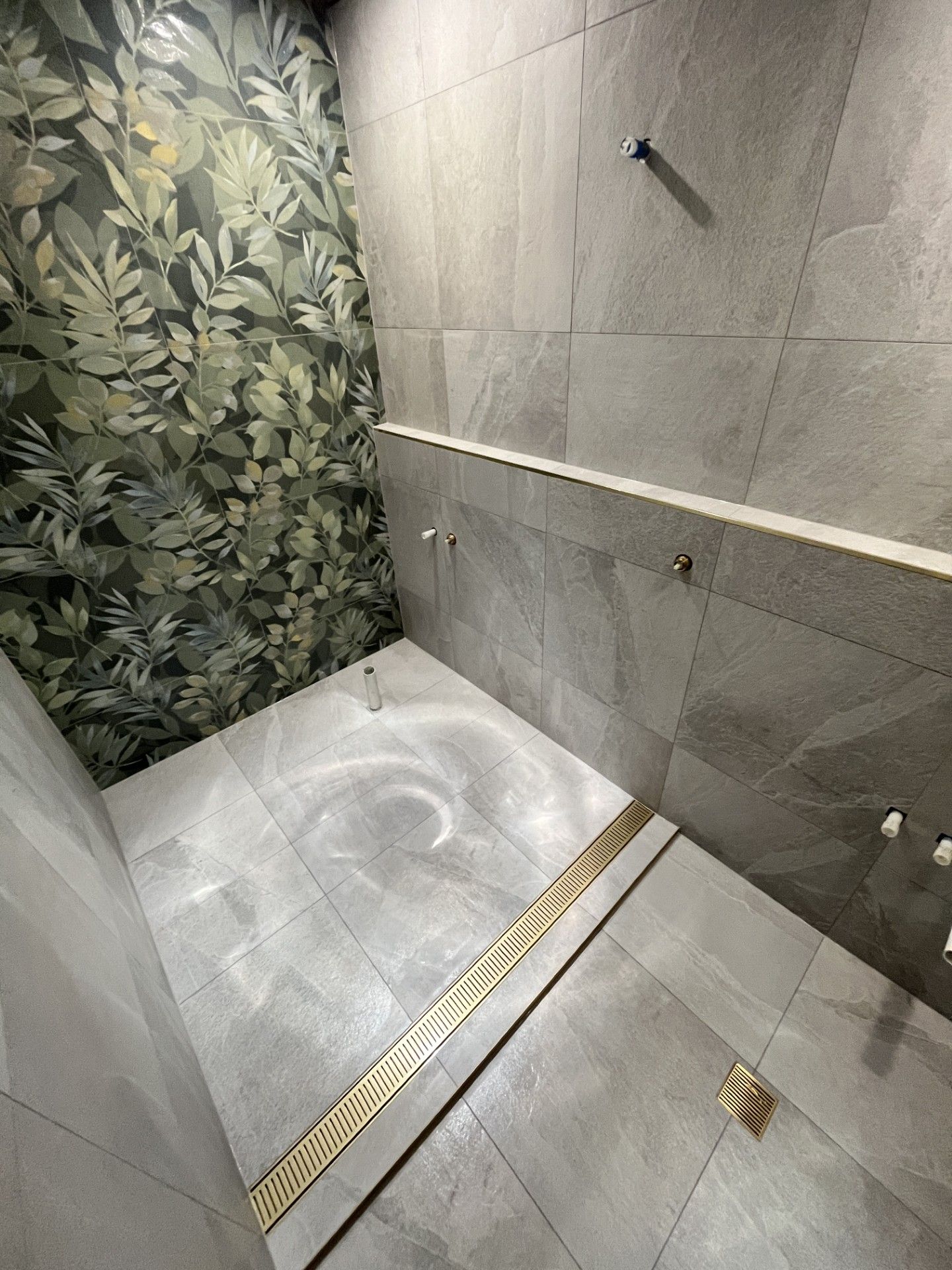 A modern walk-in shower with grey stone-textured tiles, a leafy feature wall, and a gold linear floor drain.