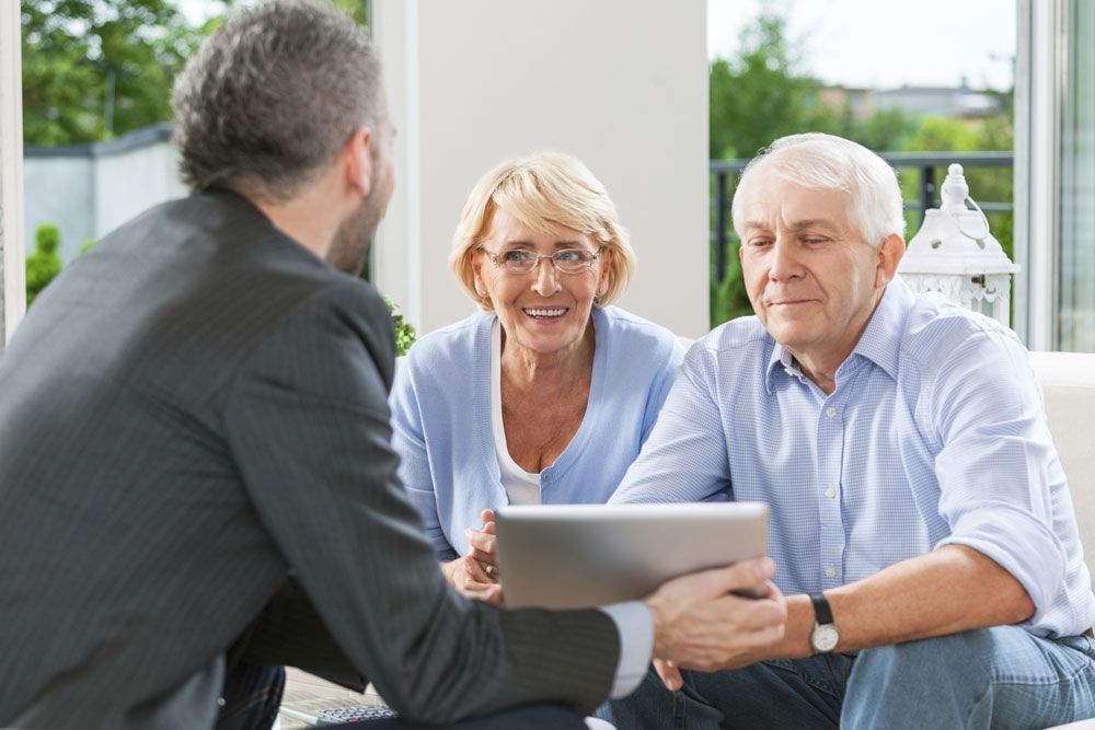 An elderly couple is sitting on a couch talking to a man while looking at a tablet.